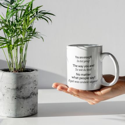 Preview: Hand hält Tasse „Du bist genug“ neben einer Pflanze